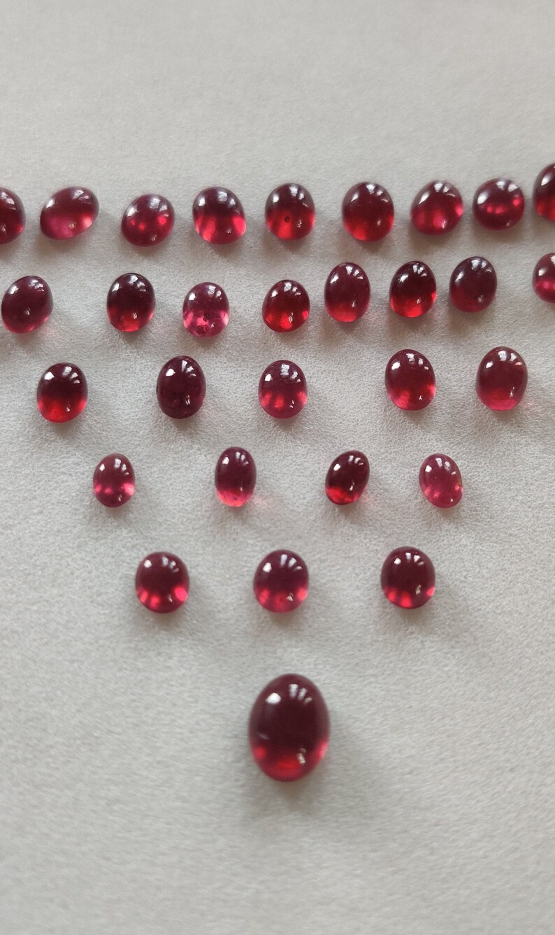 May include: A collection of 25 round, red gemstones arranged in a triangular pattern on a white surface.