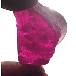 May include: A large, rough-cut, deep pink gemstone with a slight purple hue. The gemstone is translucent and has a slightly cloudy appearance.