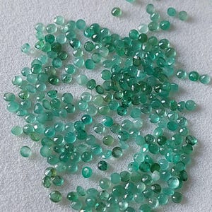 May include: A scattering of small, round emerald gemstones in various shades of green. The gemstones are spread across a light surface, with facets reflecting light. The gems appear to be of different sizes and shapes.