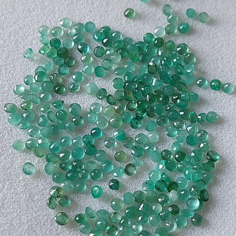 May include: A scattering of small, round emerald gemstones in various shades of green. The gemstones are spread across a light surface, with facets reflecting light. The gems appear to be of different sizes and shapes.