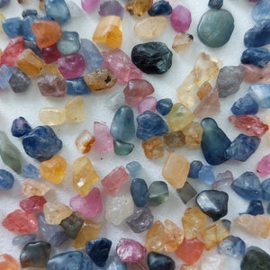 Peut inclure: Une collection de pierres précieuses facettées en vrac de différentes couleurs, notamment bleu, rose, jaune et vert. Les pierres précieuses sont éparpillées sur une surface blanche.