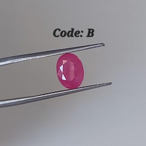 May include: A faceted, oval ruby gemstone held by tweezers. The ruby is a vibrant red colour and has a polished surface. The text "Code: B" is above the gemstone.