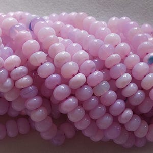 May include: Close-up of a strand of light pink gemstone beads. The beads are oval-shaped and have a polished, glossy finish. The color varies slightly, with some beads appearing more lavender. The beads are strung together, creating a flowing, organic shape.