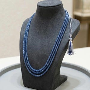 Puede incluir: Un collar de varias hebras con tres capas de cuentas azules facetadas. El collar está rematado con un adorno de borla plateada y azul que cuelga del lateral, proporcionando un contraste marcado con las hebras de cuentas azul oscuro.