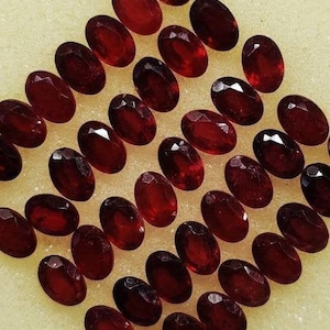 May include: A collection of 30 oval-shaped, dark red gemstones. The gemstones are arranged in a diamond pattern on a white surface.