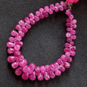 May include: A necklace of vibrant pink teardrop-shaped beads. The beads are strung together, forming a loop. The necklace is set against a dark background, highlighting the color and shape of the beads. The necklace has a tassel.
