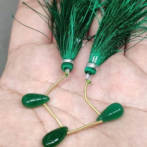 Può includere: Orecchini con nappe verde smeraldo con perline a forma di goccia. Le nappe sono attaccate ad accenti metallici argentati e collegate alle perline con filo dorato. Gli orecchini sono esposti su una mano.