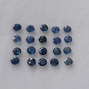 Può includere: Una collezione di venti zaffiri blu rotondi e sfaccettati. Le pietre preziose variano in tonalità di blu, da chiaro a indaco intenso, e sono disposte su una superficie bianca. Ogni pietra ha una superficie lucida e riflettente.