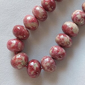 May include: A strand of oval-shaped beads in shades of pink and cream. The beads have a polished, glossy finish and are strung together on a red cord. The beads are approximately 1 cm in diameter.