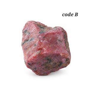 May include: A raw, unpolished ruby crystal with a deep red hue and visible dark mineral inclusions. The stone has a rough, multifaceted surface texture and irregular edges. The text code B is printed in black in the upper right corner.