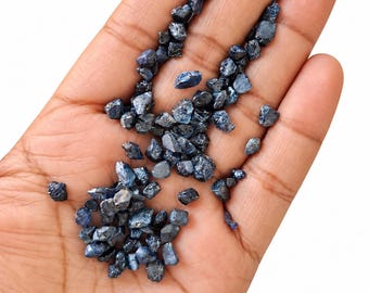 May include: A collection of small, raw, dark blue sapphire gemstones with irregular, faceted shapes and a natural, unpolished texture. These loose, deep blue stones vary in size and display a translucent to opaque appearance with reflective surfaces.