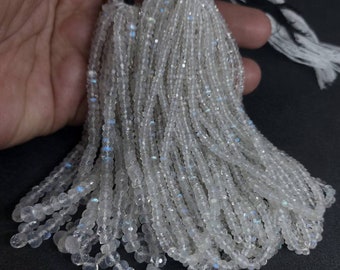 Peut inclure: Un brin de perles de pierre de lune blanche, avec un léger éclat iridescent, enfilées sur un cordon noir. Les perles sont facettées et ont une forme légèrement irrégulière.