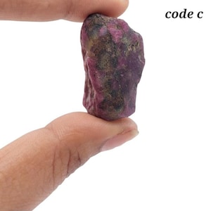 Op de afbeelding: Een ruwe, ongepolijste robijn edelsteen met een diep magenta en donkerpaars kleurenpalet. De steen heeft een onregelmatig, getextureerd oppervlak met zichtbare minerale insluitingen en natuurlijke facetten. De tekst code c is in het zwart gedrukt.