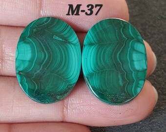 Pode incluir: Par de cabochões ovais de malaquita verde com um padrão de redemoinho. Os cabochões têm um diâmetro aproximado de 2,5 cm. A imagem inclui o texto "M-37".