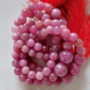 May include: A strand of pink gemstone beads, likely ruby, with a red tassel. The beads are round and smooth, and they are strung on a clear thread.