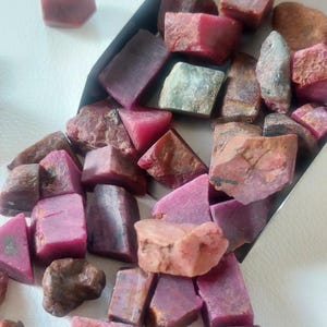 Puede incluir: Una colección de rubíes en bruto y sin pulir en varios tonos de rojo intenso, magenta y rosa. Las piedras tienen formas irregulares, con superficies rugosas y texturizadas y algunas áreas translúcidas, dispuestas dentro de una pala de metal.