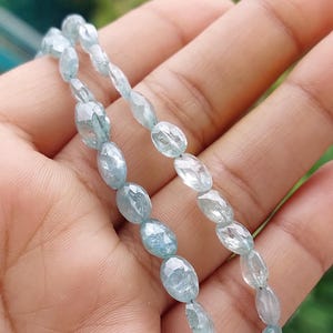 Puede incluir: Un hilo de cuentas de piedras preciosas de color azul claro, con forma ovalada y facetas. Las piedras translúcidas presentan un tono azul hielo pálido con sutiles inclusiones internas y una superficie pulida y reflectante, dispuestas en línea.