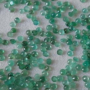 May include: A scattering of small, round emerald gemstones. The gems are various sizes and shades of green. Each gem has a faceted surface, reflecting light. The arrangement suggests a collection of loose gemstones, possibly for jewellery making. The gems are displayed on a neutral background.