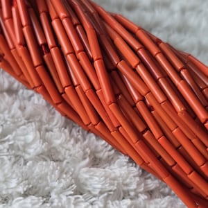 May include: A multi-strand necklace composed of numerous small, cylindrical, bright orange beads. The beads are arranged in a dense, twisted formation, creating a textured, tubular appearance with a consistent, vibrant orange color throughout the piece.