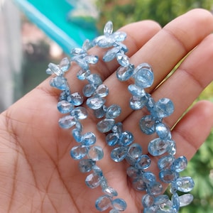 Puede incluir: Un hilo de piedras preciosas de color azul claro, talladas en forma de pera y con múltiples facetas. Cada piedra individual presenta una calidad translúcida y una superficie pulida que refleja la luz a través de sus cortes en una cadena.