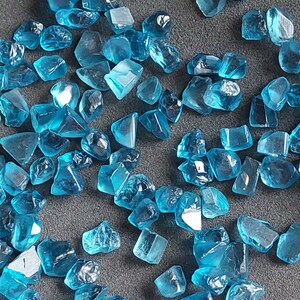 May include: A collection of loose, faceted, teal blue gemstones. The gemstones are various sizes and shapes, and have a shiny, polished finish.