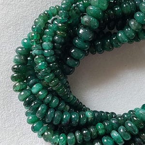 Puede incluir: Primer plano de varios hilos de cuentas de esmeralda. Las cuentas son de un color verde intenso y rico y tienen una superficie lisa y pulida. Las cuentas están ensartadas, formando un collar o pulsera.