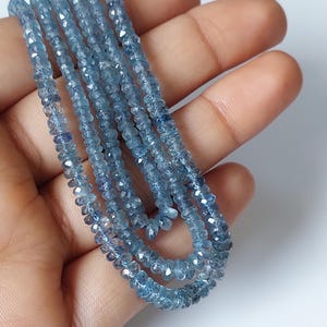 Puede incluir: Un hilo de cuentas de piedras preciosas azules, translúcidas y facetadas. Las cuentas son pequeñas, redondas y tienen una superficie tallada y reflectante que crea un aspecto brillante. El hilo está dispuesto en varios bucles.