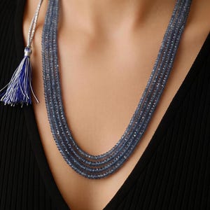 Puede incluir: Un collar de múltiples hilos con cuatro filas de pequeñas cuentas de piedras preciosas facetadas en color azul pizarra. Los hilos están asegurados con detalles metálicos plateados y terminados con una borla de hilo azul y blanco.