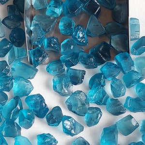 May include: A collection of loose, faceted, teal blue gemstones. The gemstones are scattered on a white surface.