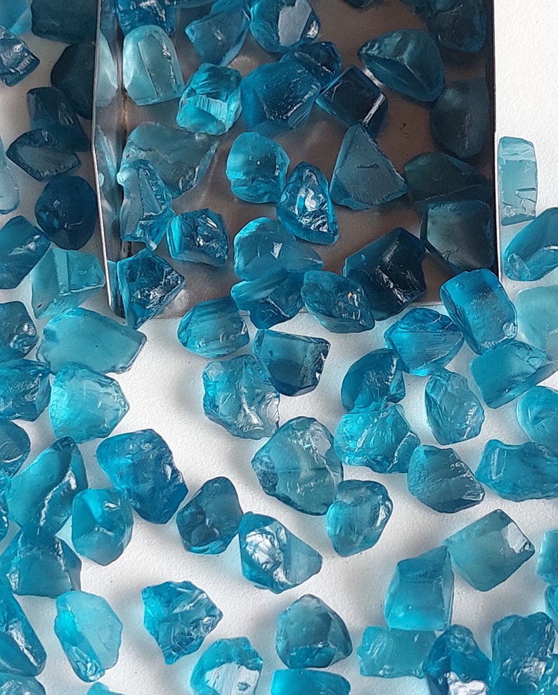 May include: A collection of loose, faceted, teal blue gemstones. The gemstones are scattered on a white surface.