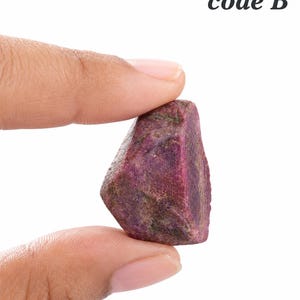 May include: A raw, uncut ruby gemstone with a deep magenta and dark red colour palette. The stone features a rough, textured surface with natural crystalline facets and irregular edges. The text code B is printed in black in the upper right corner.