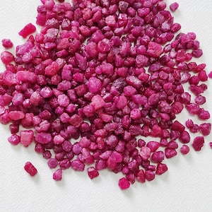 Op de afbeelding: Een verzameling kleine, ruwe, ongepolijste robijn-edelstenen in verschillende tinten dieproze en magenta. De stenen zijn onregelmatig van vorm en grootte en hebben een getextureerd, kristallijn oppervlak dat het licht over de groep weerkaatst.