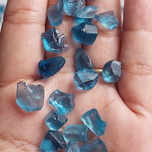 May include: A collection of blue gemstone chips, some with a slightly darker shade, scattered on a hand. The chips are irregular in shape and have a translucent appearance.