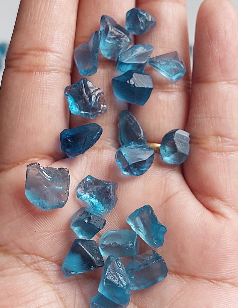 May include: A collection of blue gemstone chips, some with a slightly darker shade, scattered on a hand. The chips are irregular in shape and have a translucent appearance.