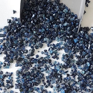 Puede incluir: Una colección de pequeñas gemas azules en bruto, con variaciones de azul marino oscuro a turquesa claro. Las gemas están dispersas sobre una superficie blanca, con una cuchara metálica parcialmente visible, sugiriendo medición o exhibición.
