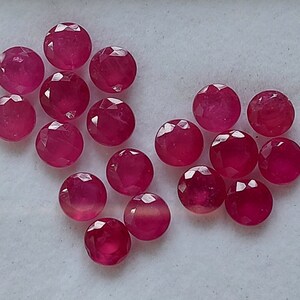 May include: A collection of 15 faceted ruby gemstones, each with a round shape and a deep red color. The gemstones are arranged in a loose, scattered pattern on a white surface.