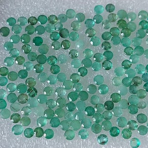 May include: A collection of small, round, faceted emerald gemstones in varying shades of green and teal. The gemstones are scattered across a white, textured surface, showcasing their brilliant cut and colour.