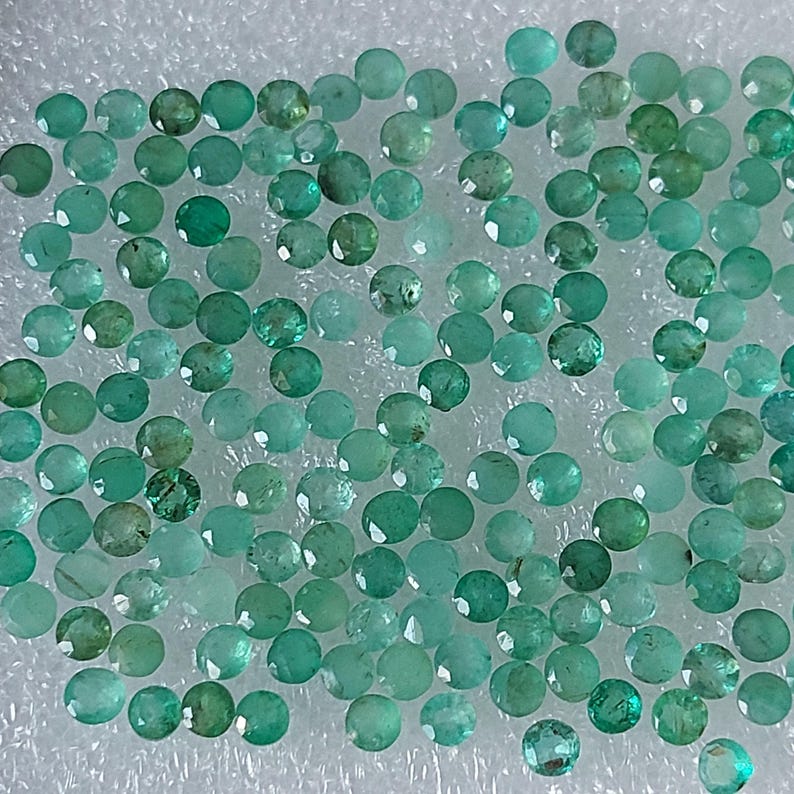 May include: A collection of small, round, faceted emerald gemstones in varying shades of green and teal. The gemstones are scattered across a white, textured surface, showcasing their brilliant cut and colour.