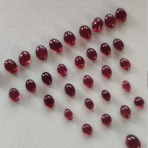 May include: A collection of oval-shaped, deep red gemstones arranged in a triangular pattern on a white surface.
