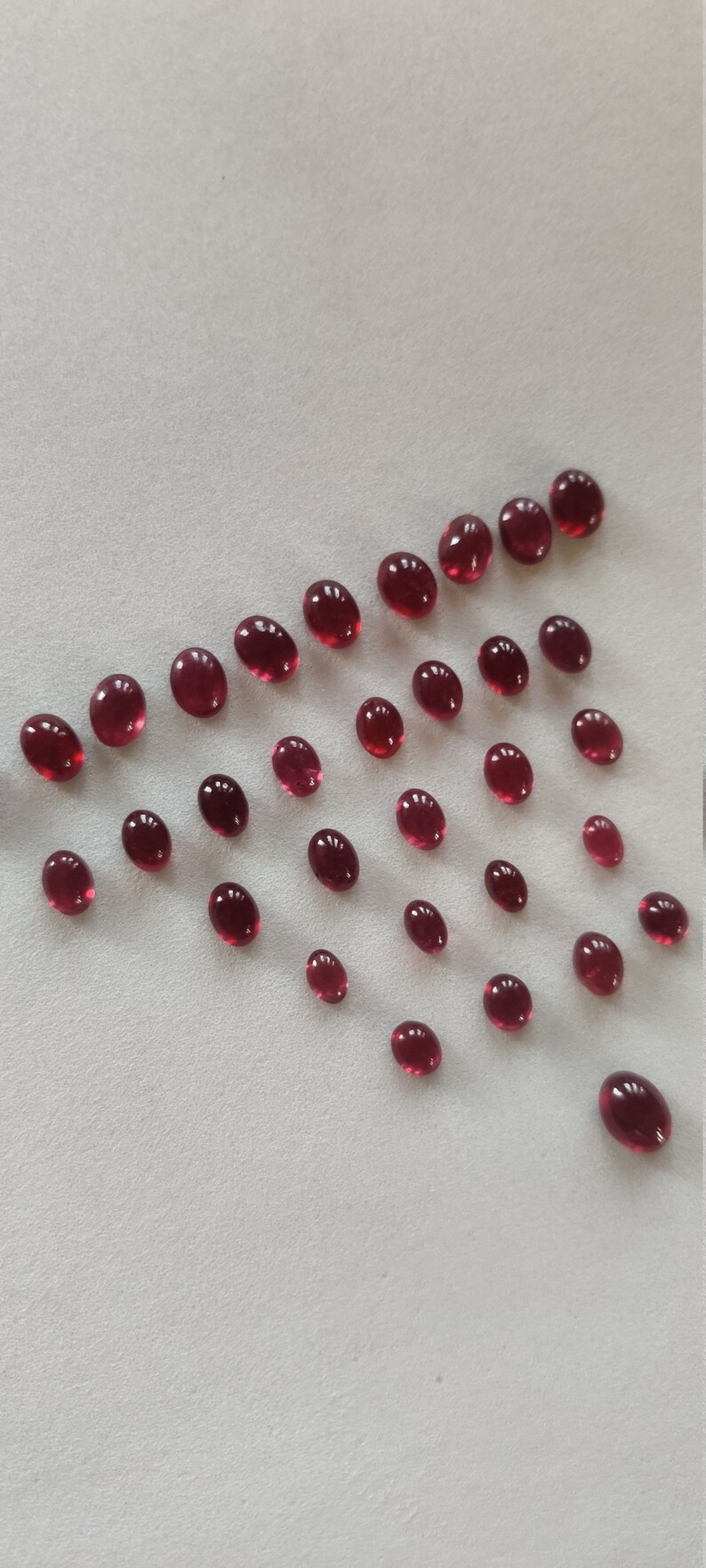May include: A collection of oval-shaped, deep red gemstones arranged in a triangular pattern on a white surface.