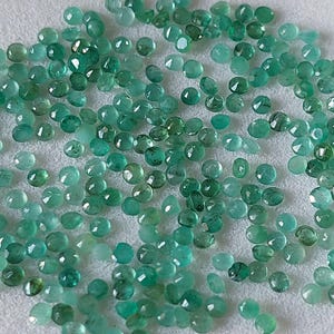 May include: A collection of small, round, faceted emerald gemstones in various shades of green. The gemstones are scattered on a white surface, with light reflecting off their polished surfaces. The gems are of various sizes.
