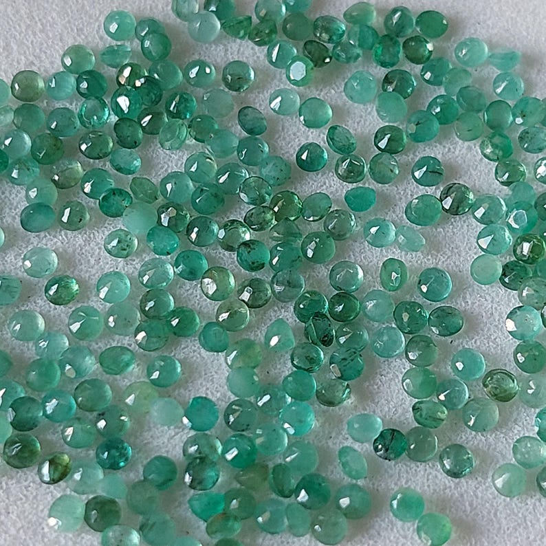 May include: A collection of small, round, faceted emerald gemstones in various shades of green. The gemstones are scattered on a white surface, with light reflecting off their polished surfaces. The gems are of various sizes.