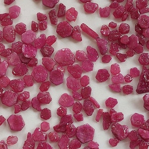 May include: A collection of loose, red ruby gemstones of various sizes and shapes. The rubies are scattered on a white surface.