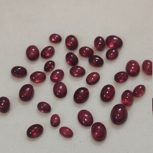 May include: A collection of oval-shaped, red gemstones. The gemstones are scattered on a white surface.
