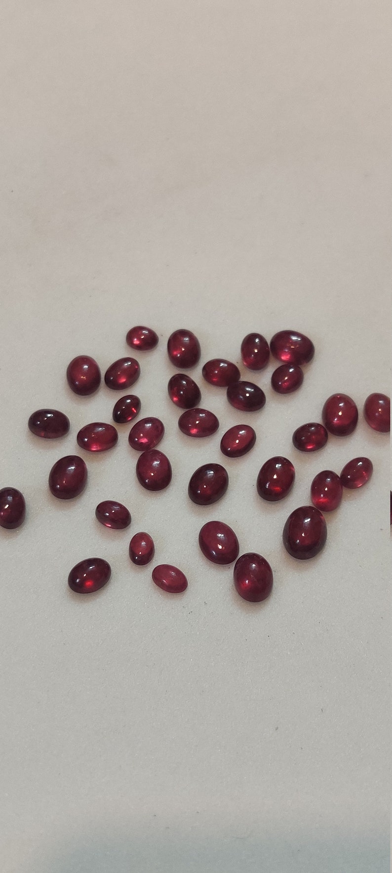 May include: A collection of oval-shaped, red gemstones. The gemstones are scattered on a white surface.
