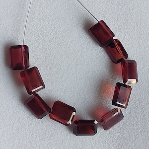 May include: A string of rectangular, faceted, dark red gemstone beads. The beads are strung on a thin, dark thread, and are evenly spaced. The gemstones have a polished, reflective surface, and are arranged in a curved line.