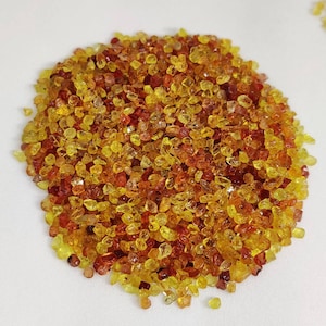 May include: A pile of small, faceted gemstones in shades of yellow, orange, and red. The gemstones are scattered across a white surface, creating a vibrant and colorful display. The gems appear to be of various sizes and shapes.
