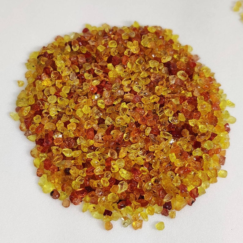 May include: A pile of small, faceted gemstones in shades of yellow, orange, and red. The gemstones are scattered across a white surface, creating a vibrant and colorful display. The gems appear to be of various sizes and shapes.