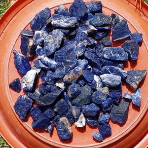 Puede incluir: Un conjunto de piedras de sodalita en bruto de varios tamaños y formas. Las piedras son principalmente de color azul oscuro, con vetas blancas y grises y algunas inclusiones marrones. Están dispuestas sobre una superficie redonda de color marrón rojizo.