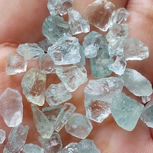 Puede incluir: Una colección de piedras preciosas de aguamarina azul claro, algunas con un ligero tono verde, en varias formas y tamaños. Las piedras preciosas son ásperas y tienen un aspecto natural sin pulir.